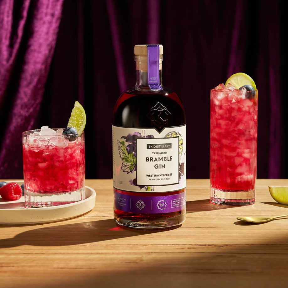 Bramble Gin bottle with two cocktails on a wooden table against a purple curtain background